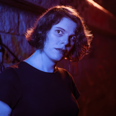 Portrait de Julia Popek. Cadrage de tête. Elle tient un couteau contre son visage. Elle est dans les ténèbres, seulement illuminée par une lumière rouge.